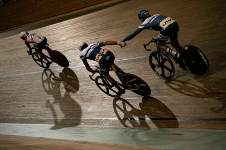 Drei Radfahrer liefern sich ein Rennen auf einem Indoor-Velodrom; ein Radfahrer streckt die Hand aus, um einen anderen anzuschieben, während ein dritter Radfahrer auf der Strecke vorausfährt, wobei alle lange Schatten auf den Holzboden werfen.