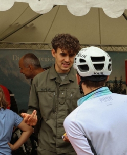 Ein Mann in einer grünen Uniform mit der Aufschrift WAFLER unterhält sich mit einem Radfahrer, der einen weißen Helm und ein hellblaues Trikot trägt, unter einem Zelt, während im Hintergrund weitere Personen und eine Kulisse zu sehen sind.