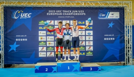 Drei männliche Radsportler stehen bei den UEC-Bahn-Europameisterschaften 2023 auf einem Podium und halten Blumen und Medaillen in der Hand. Sie lächeln vor einer blauen Kulisse mit Veranstaltungslogos und Sponsorenbannern.
