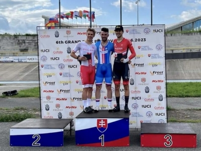 Drei männliche Radfahrer stehen auf einem Podium und halten Trophäen in der Hand. Der Radfahrer in der Mitte trägt ein blau-weißes Outfit auf dem Siegerpodest, flankiert von Radfahrern in weißen (zweiter Platz) und roten (dritter Platz) Outfits. Hinter ihnen sind Fahnen und Banner zu sehen.