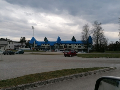 Ein Gebäude mit einem blauen Spitzdach und mehreren dreieckigen Spitzen steht hinter einem weitgehend leeren Parkplatz mit einigen Autos unter einem bewölkten Himmel. Auf der rechten Seite sind Bäume ohne Blätter zu sehen.
