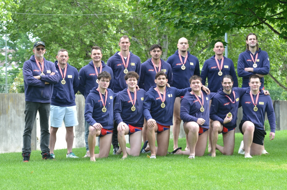 Ein Herrensportteam in passenden marineblauen Jacken und Medaillen posiert im Freien auf dem Rasen in zwei Reihen - einige kniend, andere stehend - und lächelt vor Bäumen und einer Betonmauer.