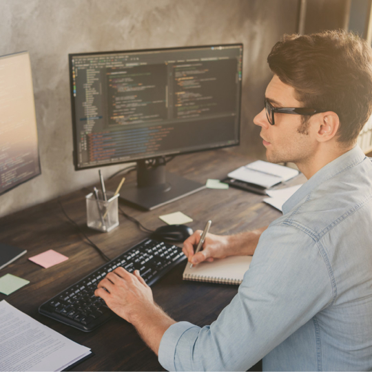 Ein Mann sitzt an einem Schreibtisch und arbeitet an einem Computer mit zwei Monitoren, die Codes anzeigen. Er tippt auf einer Tastatur, hält ein Notizbuch und einen Stift in der Hand und ist umgeben von Büromaterialien in einem modernen Arbeitsbereich.