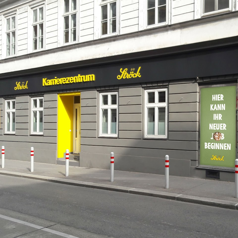 Ein graues Gebäude mit weiß gerahmten Fenstern hat eine leuchtend gelbe Tür und ein schwarzes Schild mit der Aufschrift Karrierezentrum Ströck. Auf einem Plakat neben der Tür steht Hier kann ihr neuer Weg beginnen. Weiße Poller säumen den Gehweg.
