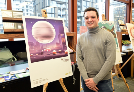 Eine Person in einem grauen Rollkragenpullover steht lächelnd neben einer Staffelei mit einem futuristischen Kunstwerk, das eine große schwebende Kugel über einer Stadtlandschaft zeigt, in einem modernen Innenraum mit großen Fenstern und weiterer Kunst im Hintergrund.