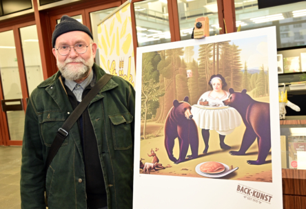 Ein Mann mit Brille und Mütze steht drinnen neben einer Staffelei, auf der ein skurriles Kunstwerk zu sehen ist, das eine Frau zeigt, die zwei Bären in einem Wald Essen serviert. Das Kunstwerk ist mit "BÄCK-KUNST" beschriftet.