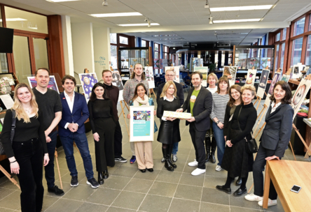 Eine Gruppe von Menschen steht in einem geräumigen, gut beleuchteten Raum zusammen, hinter ihnen sind Kunstwerke auf Staffeleien ausgestellt. Einige Gruppenmitglieder halten einen gerahmten Gegenstand und eine Urkunde in der Hand, und alle lächeln in die Kamera.