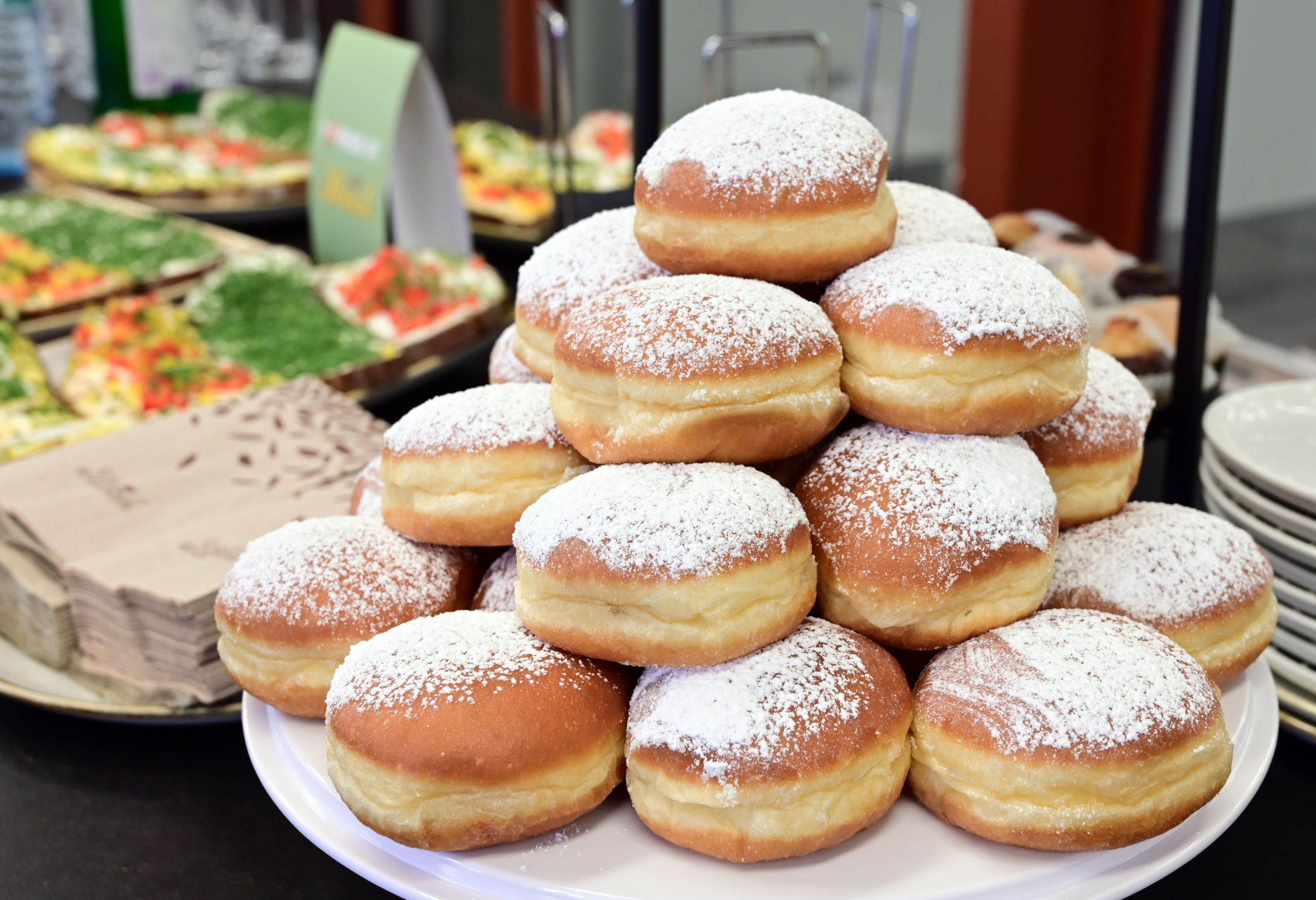 Ein pyramidenförmiger Stapel mit Puderzucker überzogener Donuts wird auf einem weißen Teller präsentiert. Im Hintergrund sind Teller, Servietten und Tabletts mit Lebensmitteln zu sehen, was auf ein Buffet oder ein Catering hinweist.