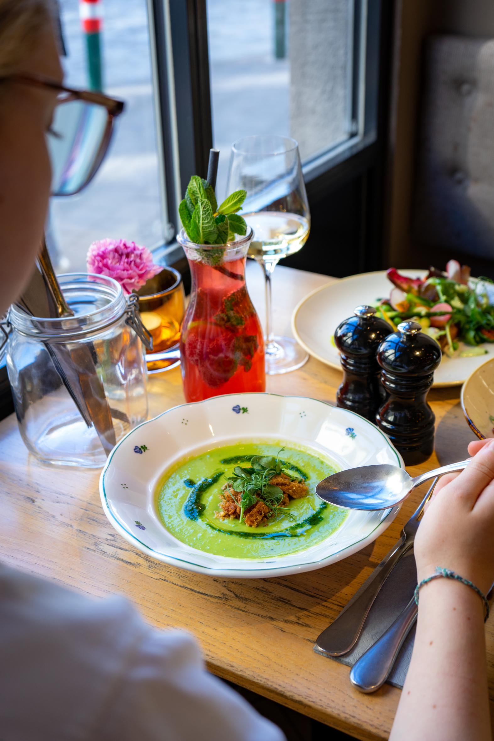 Eine Person sitzt an einem Tisch mit einer Schüssel grüner Suppe, garniert mit Kräutern, einem bunten Getränk mit Minze, einem Glas Weißwein und einem Salat. Auf dem Holztisch am Fenster stehen Salz- und Pfefferstreuer und ein Glas.