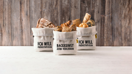 Drei mit verschiedenen Brotsorten gefüllte Stoffbeutel stehen auf einer hellen Fläche. Die Tüten sind mit deutschem Text versehen: "ICH WILL FRÜHSTÜCKEN" auf der linken und rechten Seite, und "BACKKUNST ZUM VERLIEBEN" in der Mitte. Rustikaler Holzhintergrund.