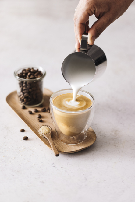 Eine Hand gießt gedämpfte Milch aus einer Metallkanne in eine Glastasse mit Kaffee. Nebenan stehen auf einem Holztablett ein Glas mit Kaffeebohnen, ein Löffel mit Zucker und einige verstreute Kaffeebohnen auf einer hellen Fläche.