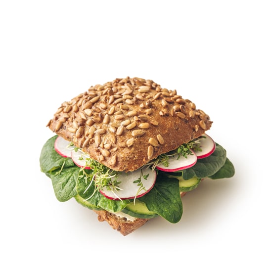 Ein quadratisches Mehrkornsandwich mit Sonnenblumenkernen obenauf, gefüllt mit Blattspinat, Avocado, Radieschenscheiben und Sprossen, vor einem weißen Hintergrund.