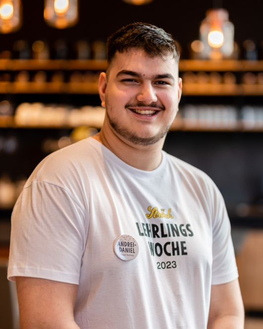 Ein junger Mann mit kurzen dunklen Haaren und einem Bart lächelt in die Kamera. Er trägt ein weißes T-Shirt mit der Aufschrift Lehrlings Woche 2023 und ein Namensschild mit der Aufschrift Andrei-Daniel. Im Hintergrund sind warme Lichter und Regale unscharf zu erkennen.