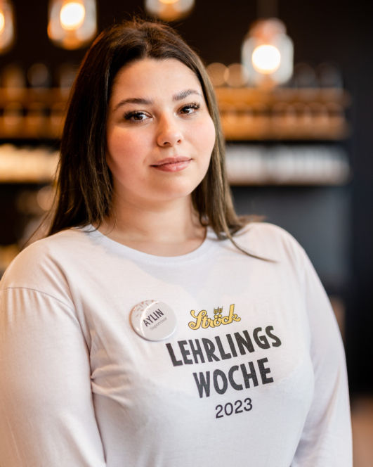 Eine junge Frau mit langen braunen Haaren trägt ein weißes Lehrlings Woche 2023-Shirt und ein Namensschild mit der Aufschrift Aylin. Sie steht in einem Innenraum mit einem sanft verschwommenen Hintergrund aus Hängelampen und Regalen.