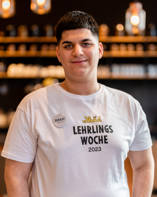 Eine junge Person mit kurzen dunklen Haaren steht lächelnd in einem Raum und trägt ein weißes Hemd mit der Aufschrift LEHRLINGS WOCHE 2023 und ein Namensschild mit der Aufschrift Berkay. Der Hintergrund ist mit Regalen und Hängelampen sanft beleuchtet.