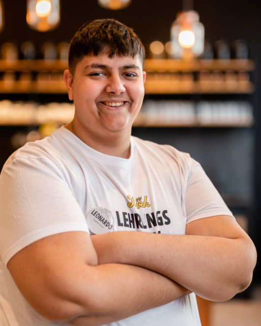 Eine junge Person mit kurzen dunklen Haaren lächelt selbstbewusst mit verschränkten Armen, trägt ein weißes T-Shirt und ein Namensschild mit der Aufschrift Leonardo und steht in einem Innenraum mit verschwommenen Lichtern und Regalen im Hintergrund.