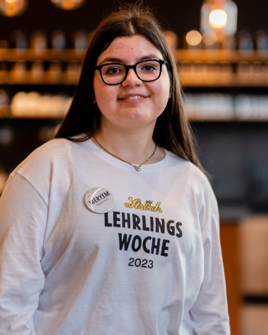 Eine junge Frau mit langen braunen Haaren und Brille lächelt in die Kamera. Sie trägt ein weißes Hemd mit der Aufschrift Lehrlings Woche 2023 und ein Namensschild. Der Hintergrund ist sanft verschwommen und warm beleuchtet.