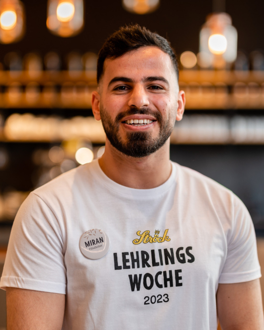 Ein lächelnder Mann mit kurzem Bart trägt ein weißes T-Shirt mit der Aufschrift Lehrlings Woche 2023 und ein Namensschild mit der Aufschrift Miran. Der Hintergrund ist sanft verschwommen mit warmen Lichtern und Regalen.