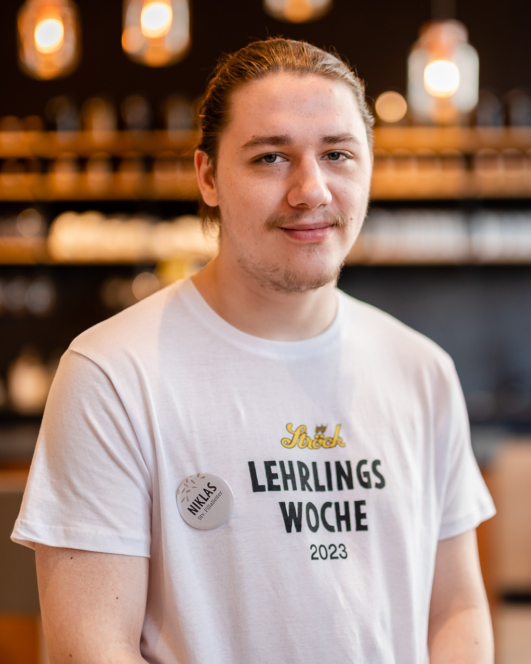 Ein junger Mann mit langen, zurückgebundenen Haaren lächelt sanft. Er trägt ein weißes T-Shirt mit der Aufschrift Lehrlings Woche 2023 und ein Namensschild mit der Aufschrift Niklas. Der Hintergrund ist unscharf und zeigt warme Lichter und Regale.