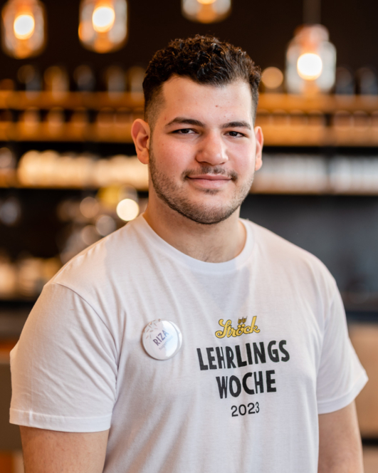 Ein junger Mann mit kurzen dunklen Haaren und gestutztem Bart steht in der Halle und lächelt. Er trägt ein weißes Lehrlings Woche 2023-T-Shirt und ein Abzeichen mit der Aufschrift Riza. Der Hintergrund ist mit warmen Lichtern sanft verschwommen.
