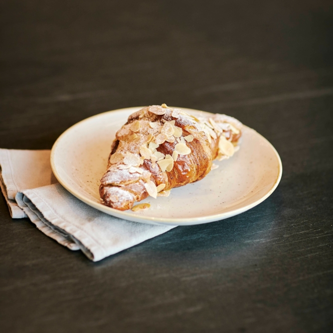 Ein Croissant mit Puderzucker und gehobelten Mandeln liegt auf einem weißen Teller, der auf einer gefalteten Serviette auf einer dunklen Fläche ruht.
