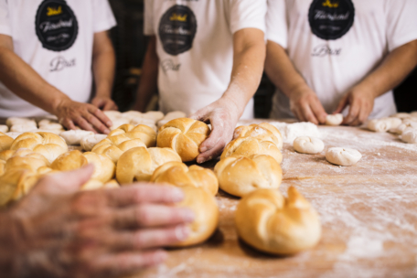 Drei Personen in weißen Hemden formen und arrangieren frisch gebackene Brötchen auf einem mit Mehl bestäubten Holztisch. Der Fokus liegt auf ihren Händen und dem goldenen Brot, im Hintergrund sind weitere Brötchen und Teig zu sehen.
