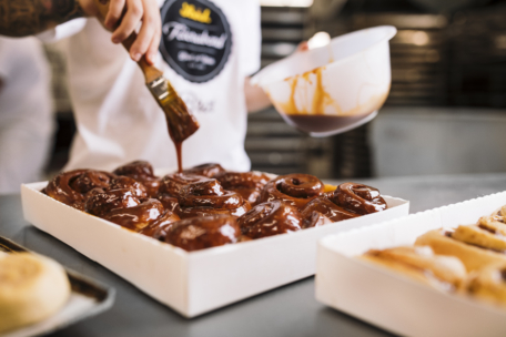 Eine Person streicht Karamellglasur auf frisch gebackene Zimtbrötchen in einem weißen Tablett, während im Hintergrund weitere unglasierte Brötchen und Küchengeräte zu sehen sind.