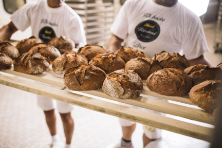 Zwei Personen in weißen Uniformen tragen in einer Bäckerei ein Holztablett mit frisch gebackenen runden Brotlaiben. Das Brot hat ein rustikales, knuspriges Aussehen.