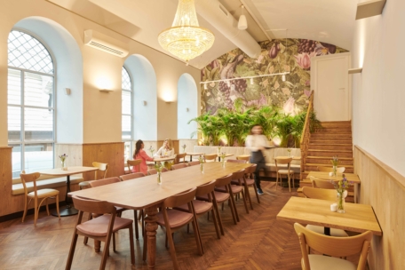 Ein gemütliches, stilvolles Café mit Holztischen und -stühlen, großen Bogenfenstern, einer Blumenwand, Zimmerpflanzen, einem Kronleuchter und einem Kellner im Vorbeigehen. Drei Personen sitzen an einem Tisch in der Nähe der Fenster und unterhalten sich.