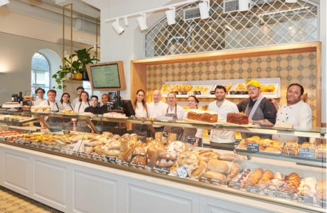 In einer hellen, modernen Bäckerei mit gefliesten Wänden und Regalen mit Backwaren steht eine Gruppe von Bäckereimitarbeitern lächelnd hinter einer Glastheke, die mit verschiedenen Brotsorten, Gebäck und Torten gefüllt ist.