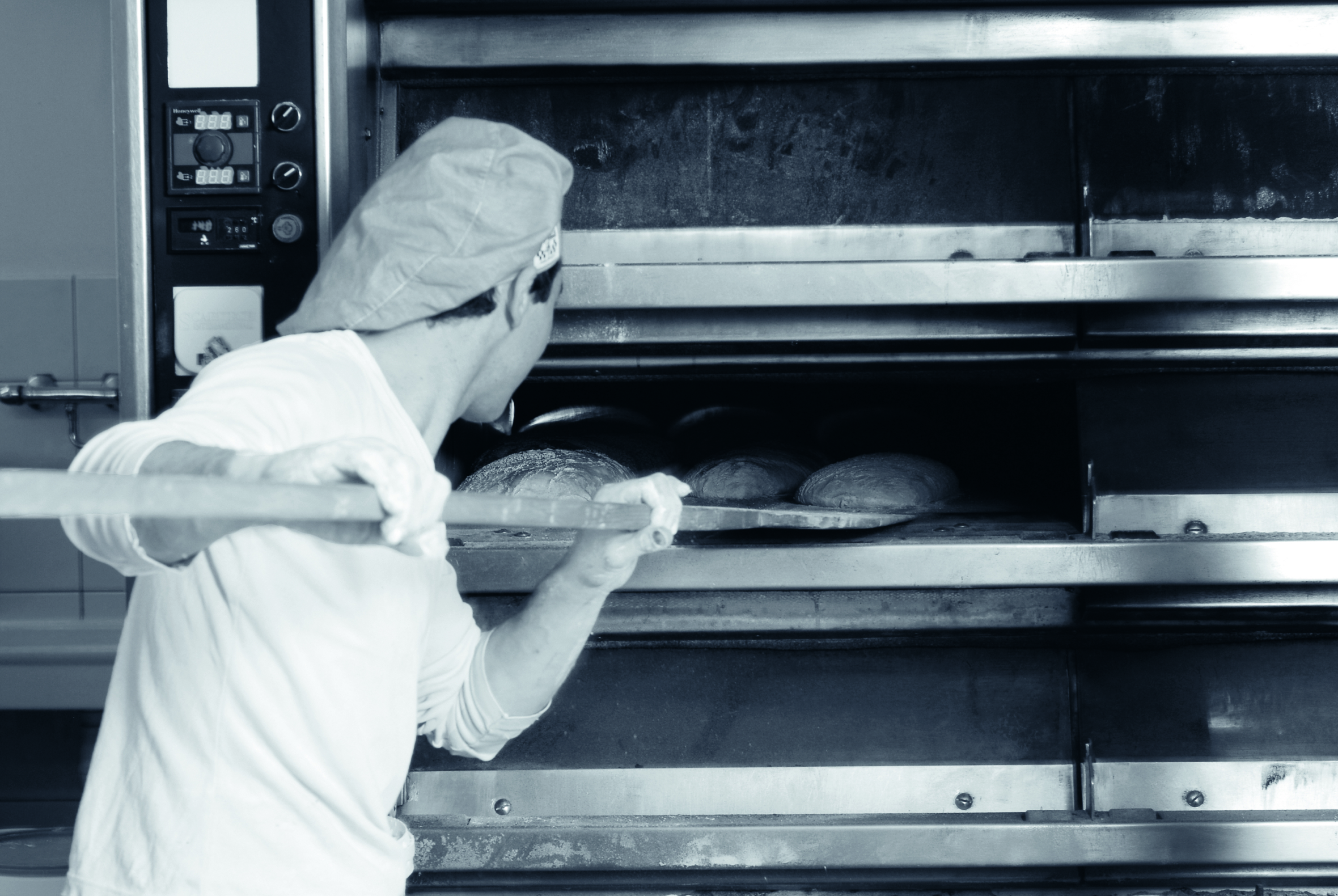 Ein Bäcker mit einer Mütze nimmt mit einem Holzschäler das frisch gebackene Brot aus einem großen gewerblichen Ofen. Die Szene wird von hinten aufgenommen, wobei der Blick auf das Brot im Ofen gerichtet ist.