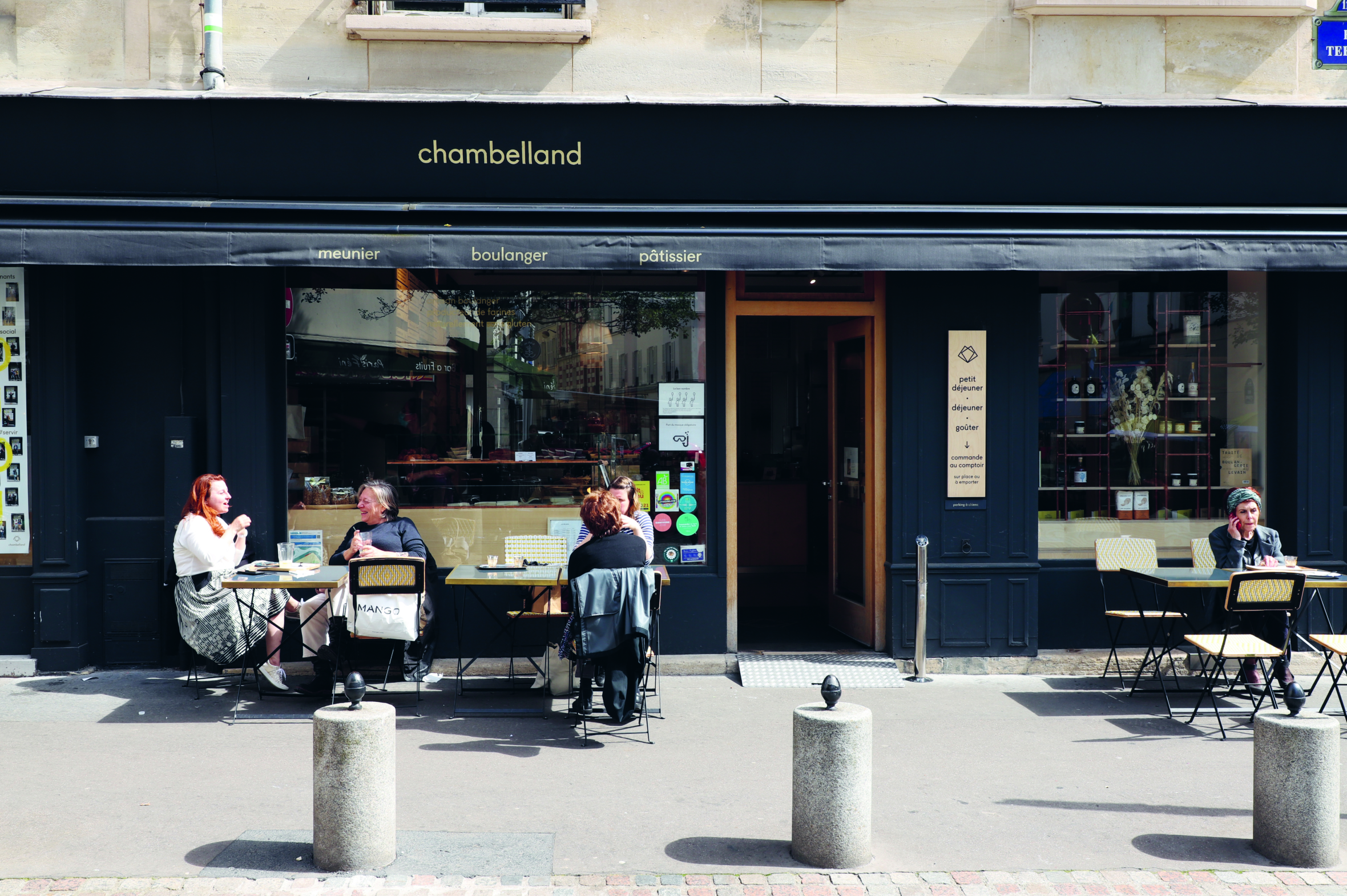 Menschen sitzen an Tischen im Freien vor der Bäckerei und dem Café Chambelland. Die Schaufensterfront hat eine schwarze Markise und große Fenster, und die Szene spielt auf einem Bürgersteig in der Stadt, auf dem Stühle und Poller zu sehen sind.