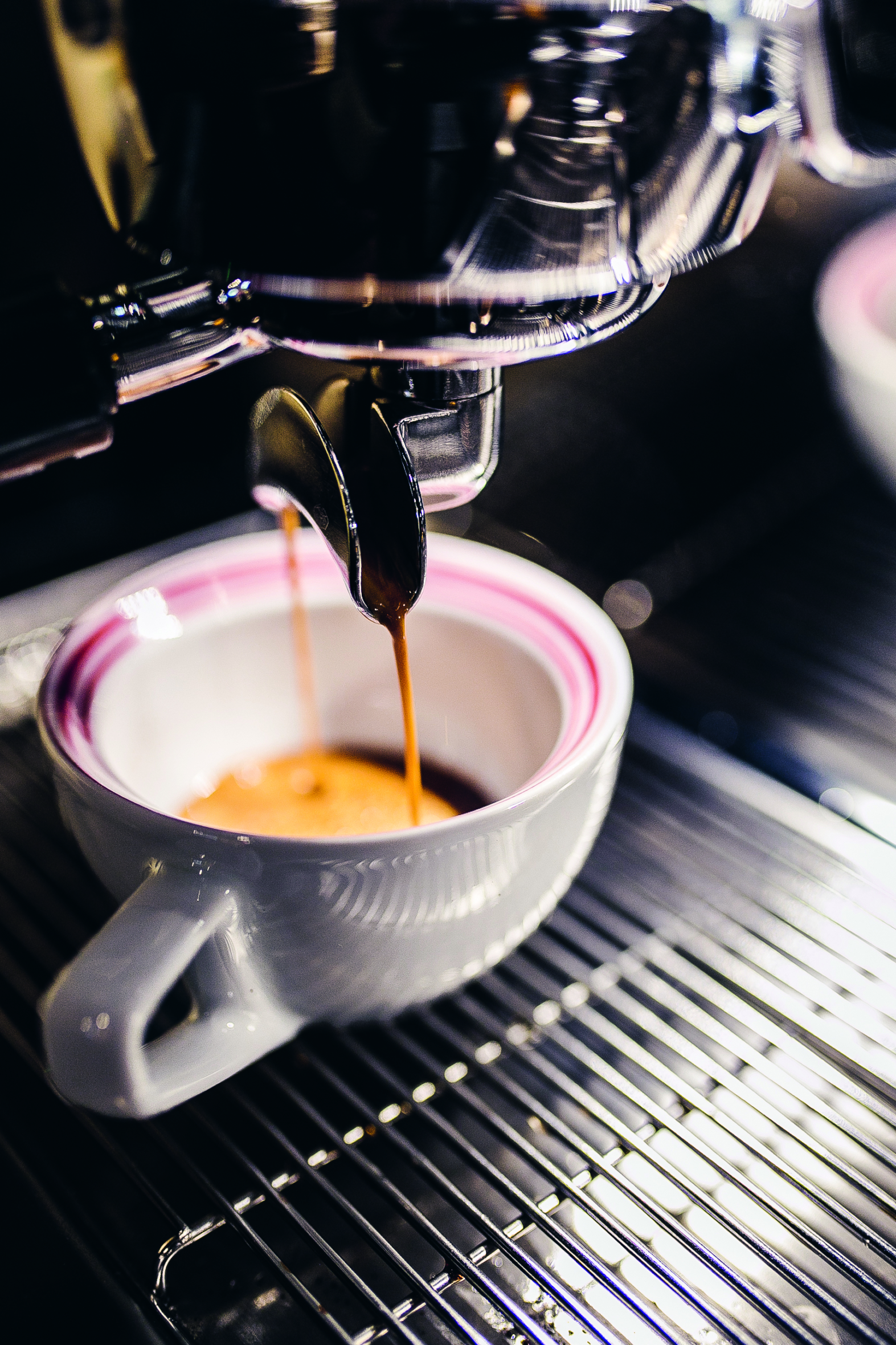 Espressomaschine, die frischen Espresso in eine weiße Tasse mit rosa Streifen gießt, die auf einem Metallgitter steht.