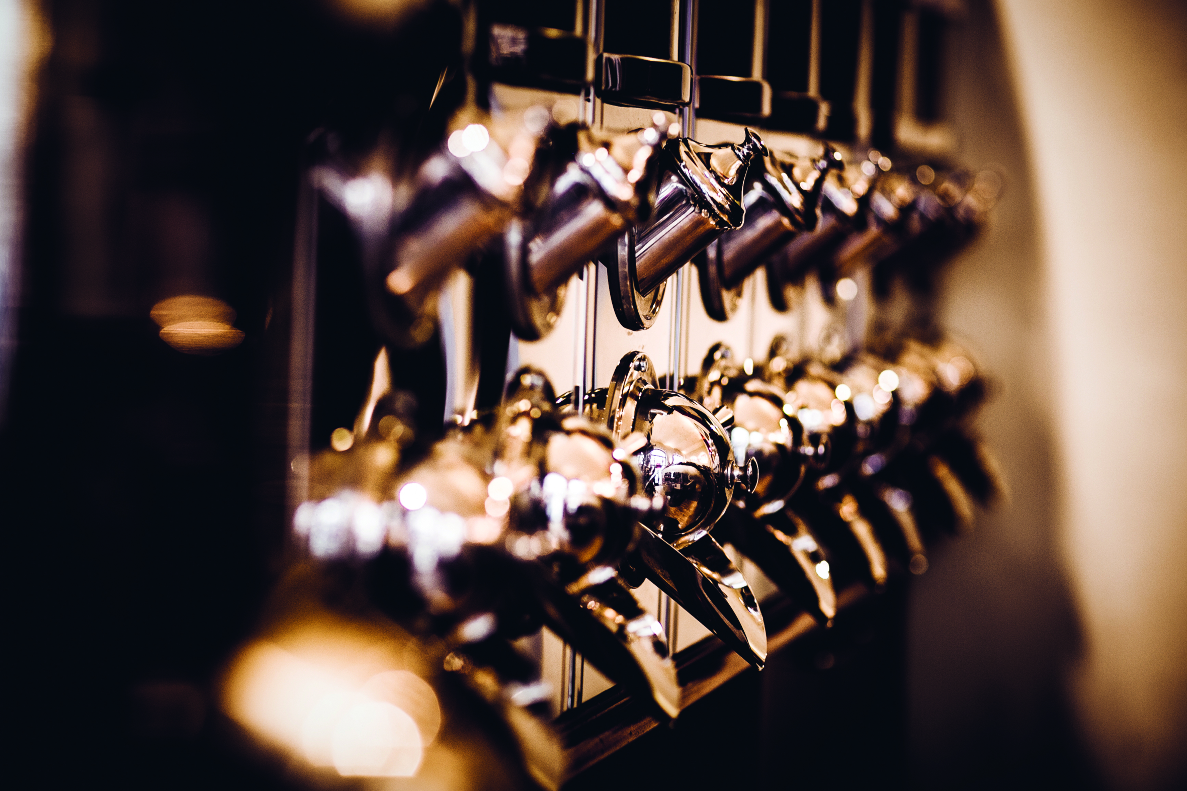 Reihen glänzender Metallzapfhähne sind auf einer Bar montiert, wobei eine warme, stimmungsvolle Beleuchtung Reflexionen auf den Oberflächen erzeugt. Das Bild konzentriert sich auf die Griffe und Ausgießer im Vordergrund, während der Hintergrund sanft verschwommen ist.