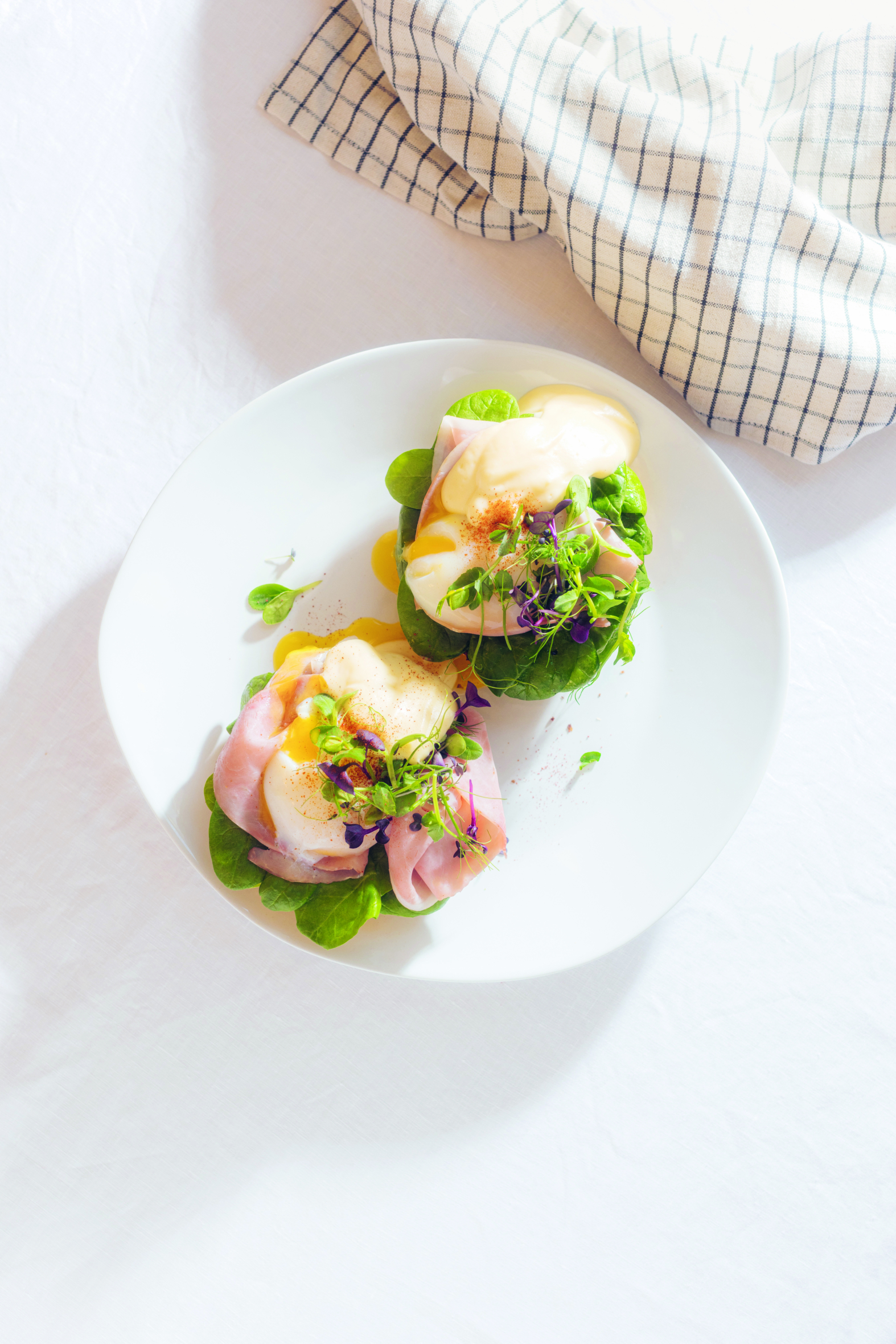 Ein weißer Teller mit zwei Portionen Eier Benedict auf Blattgemüse, jeweils belegt mit pochierten Eiern, Sauce Hollandaise, Schinken und Mikrogrün. Ein kariertes Tuch liegt im Hintergrund auf einer weißen Fläche.