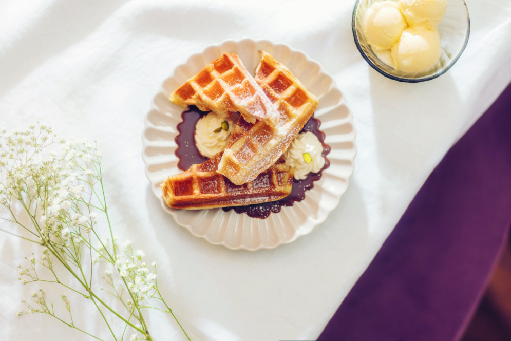 Auf einem weißen Tischtuch steht ein Teller mit drei Waffeln, die mit Puderzucker und Sahne bestreut sind, daneben eine Schale mit Vanilleeis und einige zarte weiße Blumen.
