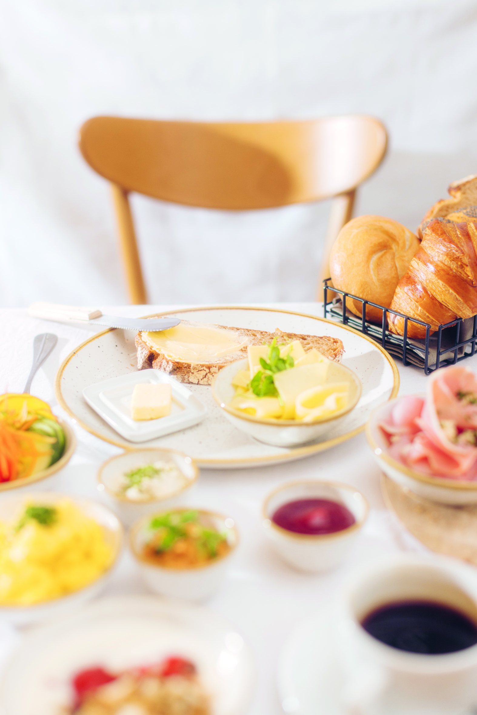 Ein gedeckter Frühstückstisch mit Brot, Butter, Käse, Gebäck, Schinkenscheiben, Gemüse, Aufstrichen, einer Tasse Kaffee und verschiedenen kleinen Speisen, mit einem Holzstuhl im Hintergrund.