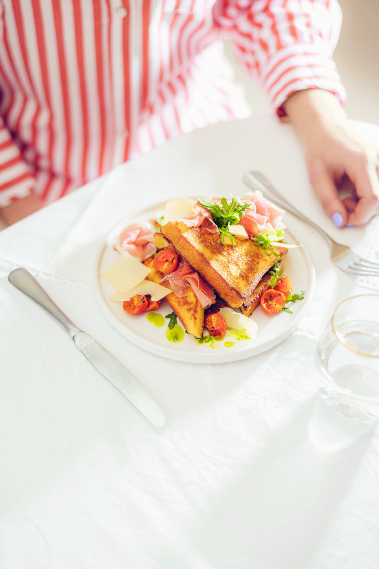Eine Person in einem rot-weiß gestreiften Hemd sitzt an einem mit Messer, Gabel und einem Glas Wasser gedeckten Tisch und ist im Begriff, einen Teller mit getoastetem Brot zu essen, das mit Tomaten, Grünzeug, Prosciutto und Käse belegt ist.