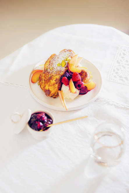 Auf einem weißen Tischtuch steht ein Teller mit French Toast, bestreut mit Puderzucker, frischen Beeren, Apfelscheiben und einer Kugel Sahne. In der Nähe stehen ein Schälchen mit Beerenmarmelade, ein Löffel und ein Glas Wasser.