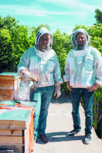 Zwei Personen in Imkerschutzanzügen stehen im Freien neben hölzernen Bienenstöcken. Eine Person hält einen Bienensmoker in der Hand, der Rauch ist sichtbar. Im Hintergrund sind grüne Bäume und ein heller Himmel zu sehen.