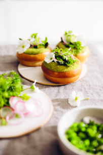 Drei runde Teigtaschen mit grüner Mousse, geschnittenen Radieschen, Kräutern und weißen essbaren Blüten sind auf kleinen Tellern angerichtet, im Vordergrund stehen frische Kräuter und Erbsen auf dem Tisch.