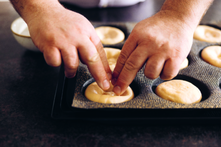 Nahaufnahme von Händen, die mit den Fingern Teig in eine Muffinform drücken, während sie den Teig auf einer dunklen Arbeitsfläche vorbereiten.