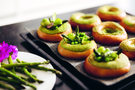 Auf der Theke steht ein Tablett mit runden Brötchen, die mit grünem Aufstrich, Spargel, Erbsen und frischen Kräutern belegt sind. Ein Teller mit Spargelstangen und einer lila Blume steht daneben.