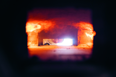 Blick aus dem Inneren eines Backofens mit Flammen an der Decke. Eine Person ist teilweise außerhalb der Ofenöffnung zu sehen, die in den Ofen hineinschaut und vom warmen Schein des Feuers angestrahlt wird.