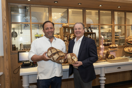 Zwei Männer lächeln in einer Bäckerei, jeder hält einen großen Laib handwerklich hergestellten Brotes in der Hand. Hinter ihnen sind Regale mit weiterem Brot zu sehen, sowie eine Theke und eine Holzauslage.