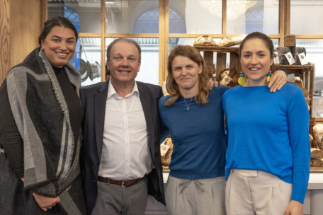 Vier Personen stehen dicht beieinander, lächeln in die Kamera und stehen vor Regalen, die mit verschiedenen Brotlaiben gefüllt sind. Die Gruppe besteht aus drei Frauen und einem Mann, die in eleganter Freizeitkleidung gekleidet sind. Die Umgebung scheint eine Bäckerei oder ein Café zu sein.