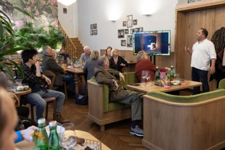 Eine Gruppe von Menschen sitzt in einem gemütlichen Restaurant um Tische herum und hört einem Mann zu, der vor einem Fernsehbildschirm steht und präsentiert; die Atmosphäre ist warm und engagiert.