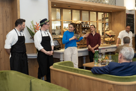 Eine Frau im blauen Pullover spricht in einem gemütlichen Restaurant zu den sitzenden Gästen, während zwei Köche mit Schürzen und Mützen und zwei Männer in der Nähe stehen. Im Hintergrund sind Regale mit Brot und Holzdekor zu sehen.