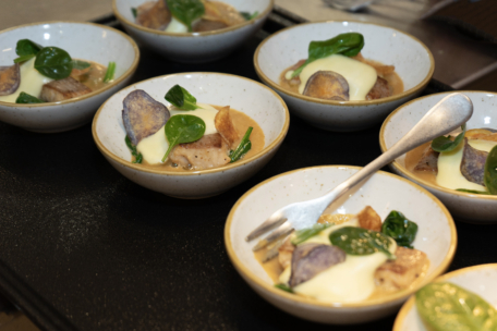 Mehrere kleine Schalen mit Gourmetgerichten, die jeweils Grünzeug, lila Kartoffelchips, Fisch- oder Fleischstücke und eine cremige Soße enthalten. In einer Schale liegt eine Metallgabel. Die Gerichte sind auf einem dunklen Serviertablett angeordnet.