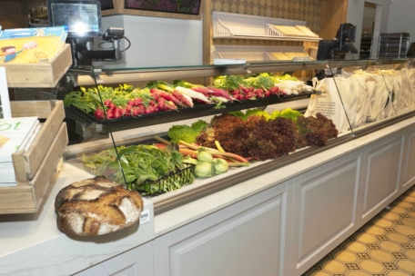 Auf einer Markttheke wird frisches Gemüse wie Radieschen, Salat und Grünzeug angeboten, und im Vordergrund steht ein runder Laib rustikales Brot. Im Hintergrund sind weiße Papiertüten und leere Brotregale zu sehen.